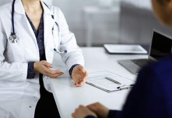 Unknown woman-doctor is talking to her patient about her diagnosis, while sitting together at the desk in the cabinet in a clinic. Physician at working place, close-up. Perfect medical service in a