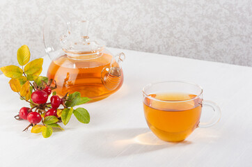 Cup of rose hip tea with teapot and autumn ripe dog rose branch on white wood table with copy space.