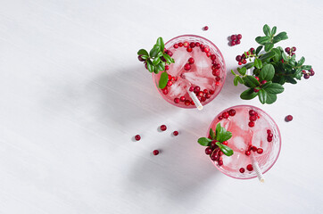 Pink cold berry cocktail with lingonberry, ice and green leaves in sunbeam on white wooden table, copy space, top view.