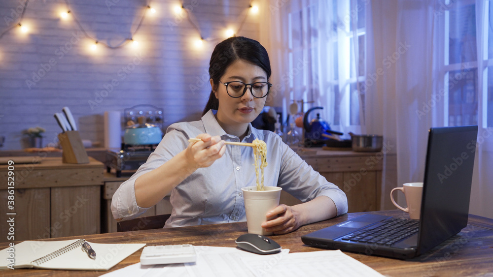 Wall mural asian female entrepreneur working in home kitchen late in night and eating instant noodle sitting at