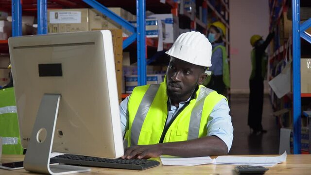 african warehouse manager sitting at desk using computer check inventory items online at distribution warehouse. black man worker  in Hard Hat typing keyboard entering data document  in workplace