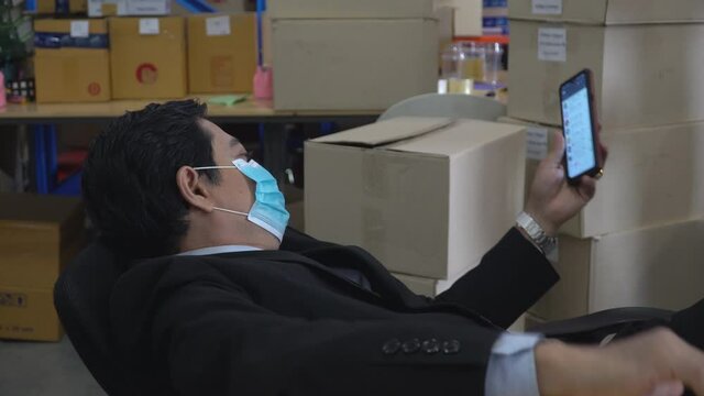 Lazy asian warehouse manager wearing face protective medical mask lying on chair using mobile phone   instead of working at storehouse