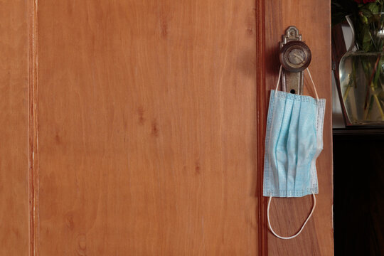 A Blue Face Mask Hanging From A Door Knob On A Wooden Door Next To A Table, Illustrating The Intrusion Of Face Masks In Our Lives During The Pandemic With Copy Space