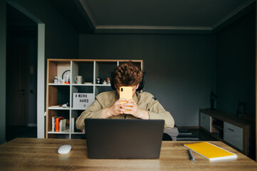Remote worker at home sits at a desk with a laptop, holds a smartphone and hides. Freelancer with a smartphone in hand uses the Internet while working on a laptop.