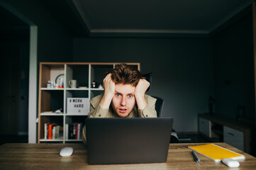 Sad young freelancer man sitting at work at home in bedroom with laptop and looking at camera with...