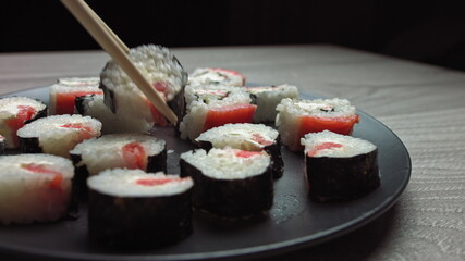 Smooth hit on a plate with Japanese food. Female hand with chopsticks takes sushi from a plate.Sushi rolls japanese food rotated over black background. California Sushi roll with tuna, vegetables