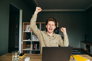 Portrait of a joyful young freelancer man sitting at a laptop at work in a cozy room and looking at...