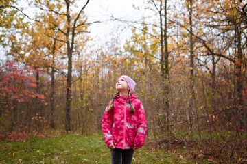 Little girl in pink jacket and knit cap standing in autumn forest