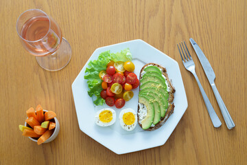 Eggs and avocado toast on white octagonal plate, cup of carrots and glass of rose wine