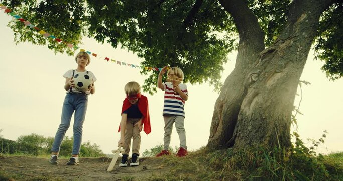 Portrait shot of the three Caucasian teen little boys playing at the big tree in summer- one in the red mask and mantle of superhero with a sword like a knight, one with a football ball and one in the