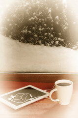 on the windowsill of a snow-covered window in winter there is a cup of black coffee and a tablet with headphones