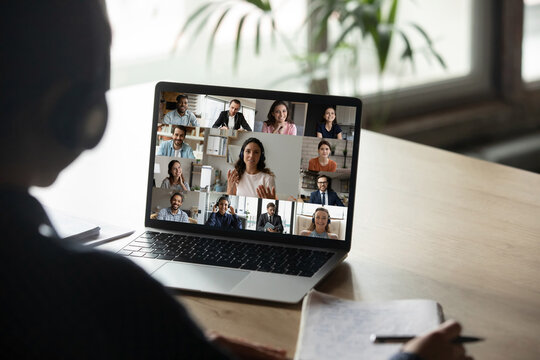 Rear view student wearing headphones engaged in online conference, using laptop, writing notes, sitting at desk, intern studying online, internet negotiations with diverse colleagues, online meeting
