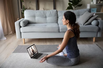 Tuinposter Sporty motivated woman wearing sportswear practicing yoga, sitting in lotus pose on warm floor in living room, using laptop, watching webinar or doing exercise with personal trainer at home © fizkes