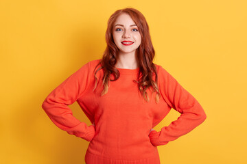 Happy woman with charming smile posing isolated over yellow background. posing with hands on hips, expressing happiness, looks at camera, wearing orange sweater.