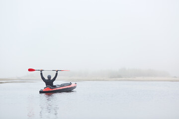 Male padding in river padding, sitting in canoe with raising paddle, enjoying water sport and beautiful nature, wearing black jacket and gray cap.