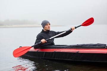 Man paddles red and black kayak with paddle in middle of river or lake in fall season. Autumn and winter kayaking, concentrated man rowing boat in foggy day. © sementsova321