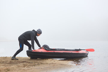 Young sportsman being ready rowing canoe, pushing boat into water, kayaker in warm clothes, padding...