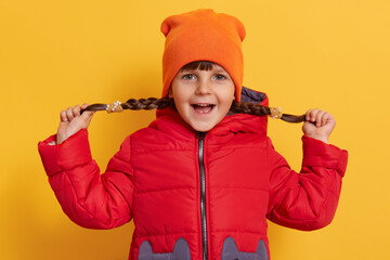 Little girl pulling her pigtails to sides, looking at camera with excited facial expression, keeps mouth opened, wearing warm clothing, standing isolated over yellow background.