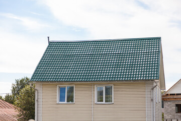 The roof of an old wooden house covered with sheets of green metal tiles on a background of green coniferous trees on a summer day. Business selling building materials or helping low-income families.
