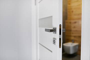 Half opened door into the cozy home interior. Modern style door handle on natural wooden white door. Chrome door handle