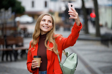 Cheerful young european woman in coat taking selfie on smartphone in the city