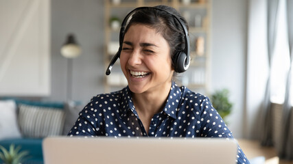 Cheerful Indian female wear headset with microphone sit in front of laptop laughing during video...