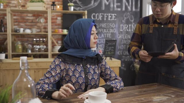 handsome asian chinese male waiter holding billing receipt folder and showing to muslim woman customer at wood table in cafe bar.