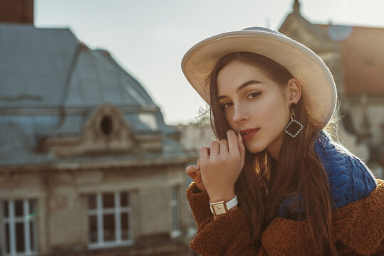 Outdoor fashion portrait of young elegant fashionable brunette woman, model wearing stylish white hat, wrist watch, brown faux fur coat, posing at sunset, in European city. Copy empty space for text
