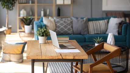 Fototapeta na wymiar Sunlight illuminates living room workplace home office interior for comfort productive work, brown and blue colours. On wooden table laptop comfy couch with cushions on background, workday end concept