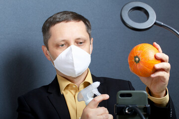 A man in a suit without a tie. Medical mask on the face. Demonstrates a pumpkin and a spray bottle with an antiseptic on camera. In front of him is a stand for a smartphone. Video communication.