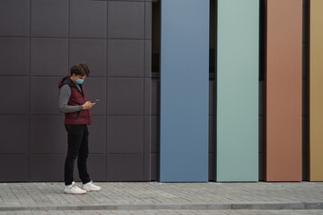 A young man walks down the street in a mask with a phone in his hands. Lifestyle, virus protection concept