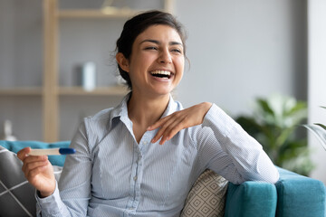 Happy Indian 30s female sit on sofa hold pregnancy positive two stripes test laughing enjoy news...