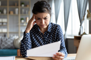 Indian woman sit at desk hold document read paper letter feels disappointed shocked by bad news....