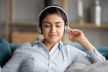 Peaceful attractive 25s Indian girl listens favourite tune through modern wireless headphones...