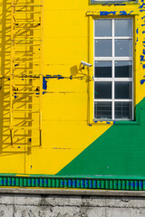 A fire escape next to a window on a building façade painted with peeling yellow and green paint.