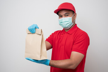 Fototapeta na wymiar Young asian delivery man in red uniform wearing protection mask and medical rubber gloves giving the paper bag to customer on isolated white background. Safety deliver concept.