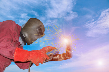 Low angle view of veiled welder welding with spark light in red unifrom with hat uniform and sunglasses on metal steel pipe of construction site on blue sky background.