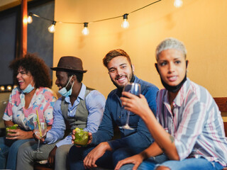 Happy multiracial friends laughing with cocktails in hands while wearing surgical face mask under chin - Celebrate at home outdoor - Coronavirus lifestyle