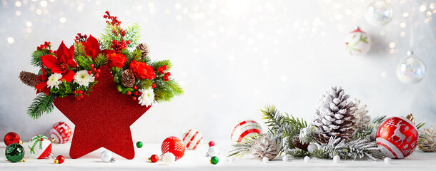 Festive winter flower arrangement in vase of red star shape and Christmas baubles on table. Christmas flower composition for holiday.