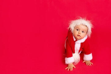 a small child of six months in a Santa costume crawls on a red isolated photophone and looks at the side, a place for text, the concept of the new year and holiday