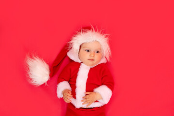 a small child of six months in a Santa costume lies on a red isolated photophone, a place for text, the concept of the new year and holiday