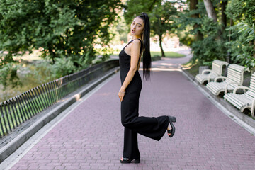 Outdoor portrait of beautiful sexy mixed raced Asian woman in black overalls, posing at park alley during sunset walk, with one leg raised up. Pretty brunette girl in elegant clothes at park