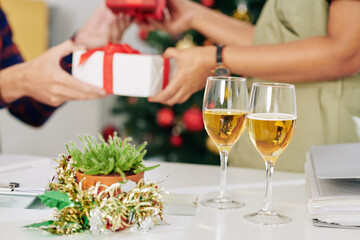 Two glasses of champagne on office table, coworkers exchanging Christmas presents in background