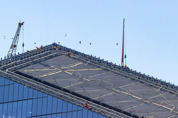 Fototapeta premium Russia,Saint Petersburg,08.09.2020.Team of workers, dressed in signal vests and carbines, works on the roof of a skyscraper. Lifting crane installed nearby.Concept of violation of safety regulations.
