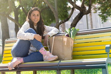 Portrait of young woman dressed casual, sitting on a bench resting after go grocery shopping. Woman with a ecological bag with a healthy purchase of fruit and vegetables