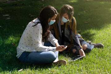 Couple of friends with medical mask, sitting in the park using their smartphone and tablet during the coronavirus pandemic