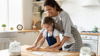Smiling loving mother and little daughter wearing aprons rolling out dough together, caring mum...