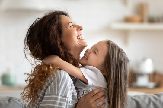 Close up side view overjoyed smiling young mother and daughter hugging and laughing, enjoying tender moment, happy mum and adorable preschool girl kid cuddling, having fun together at home