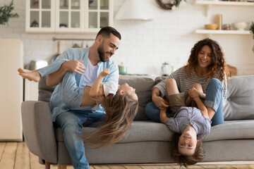 Happy young parents playing with two little kids at home, sitting on cozy couch, overjoyed mother and father holding adorable son and daughter upside down, tickling, family enjoying leisure time