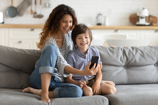 Smiling young mother and little son using phone together, hugging, sitting on cozy couch at home, happy mum with adorable kid boy taking selfie, shooting vlog, enjoying leisure time with gadget